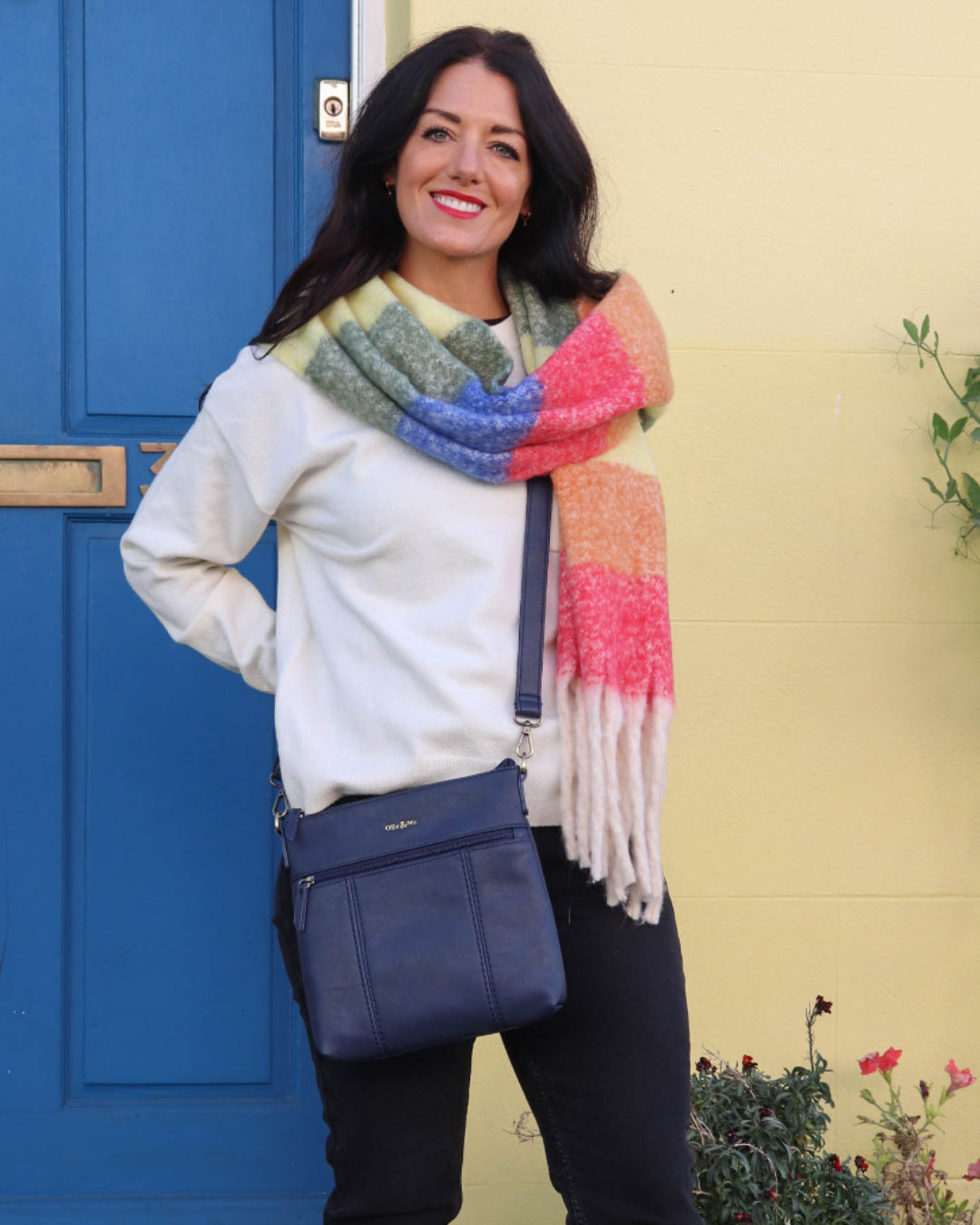 Woman wearing a colorful scarf and blue bag standing in front of a blue door and yellow wall.