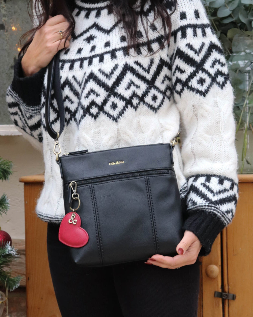 Person wearing a black and white patterned sweater holding a black Eddie Crossbody with a red heart charm.